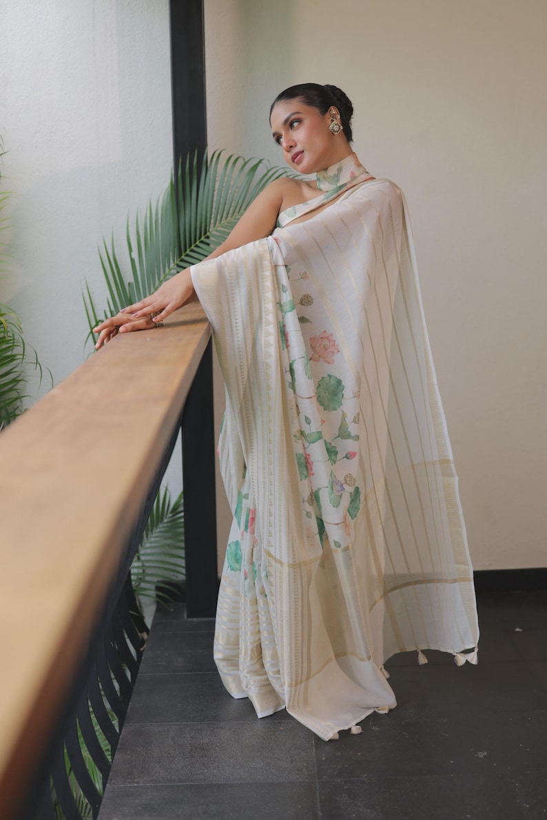 May include: An off-white saree featuring a delicate floral print with pink lotus flowers and green leaves. The fabric is adorned with vertical gold metallic stripes and a wide, textured gold border. The garment includes a matching high-neck collar.