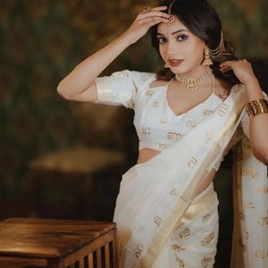 Puede incluir: Una mujer con un sari blanco tradicional con detalles dorados y escritura. Lleva una blusa a juego y joyas de oro, incluyendo un collar, pendientes y un tocado. El conjunto es elegante y culturalmente significativo.