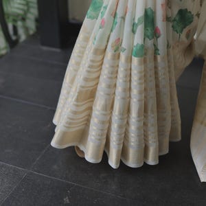 May include: A white saree featuring a floral print of pink lotus flowers and green leaves. The lower section of the fabric is adorned with horizontal bands of metallic gold thread, creating a shimmering, textured border along the pleated hemline.