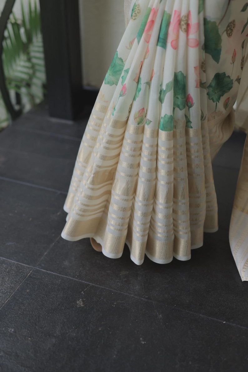 May include: A white saree featuring a floral print of pink lotus flowers and green leaves. The lower section of the fabric is adorned with horizontal bands of metallic gold thread, creating a shimmering, textured border along the pleated hemline.