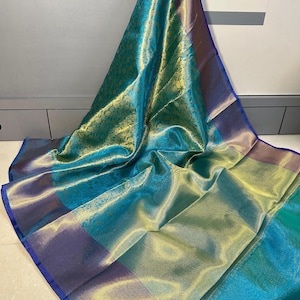 Puede incluir: Un sari brillante en color turquesa y dorado con un borde azul. La tela tiene un sutil estampado floral y un brillo metálico. El sari está drapeado, mostrando su textura fluida y variaciones de color.