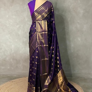 K&ouml;nnte beinhalten: Ein tiefvioletter Sari mit einem breiten goldenen Rand, der mit komplizierten Streifen- und geometrischen Mustern versehen ist. Der Stoff ist durchgehend mit kleinen goldenen Kreismotiven verziert. Eine violette &auml;rmellose Bluse ist am Torso der Schneiderpuppe zu sehen.