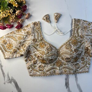May include: A white blouse featuring intricate gold and beige floral embroidery throughout the fabric. The garment has short sleeves, a sweetheart neckline, and a back closure with two white fabric tassels attached to thin white strings.