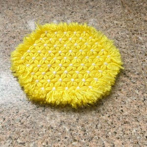 May include: A yellow crocheted pot holder with a white flower pattern and a fringe border.