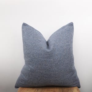 Op de afbeelding: Een groot, vierkant, denimblauw sierkussen met een textuurstof. Het kussen ligt op een houten oppervlak tegen een witte achtergrond. Het kussen heeft een zachte, uitnodigende uitstraling, geschikt voor interieurdecoratie.