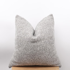 May include: A square, textured cushion in shades of grey. The cushion has a soft, nubby appearance and is placed on a wooden surface. The neutral colour palette makes it suitable for various home decor styles.