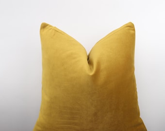 May include: A square, mustard-yellow decorative pillow. The pillow is made of a soft, velvety fabric and is placed on a wooden surface. The pillow has a simple, elegant design and is a solid color.