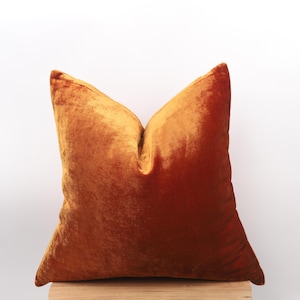 May include: A square, burnt orange velvet pillow with a soft, textured appearance. The pillow has a classic, pointed shape and is likely used for decorative purposes on a bed or sofa. The color is a warm, inviting shade.