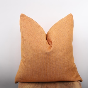 Könnte beinhalten: Ein quadratisches Kissen in einem warmen, gold-orangenen Farbton. Das Kissen hat eine strukturierte Oberfläche mit sichtbaren Webdetails, die ihm ein weiches Aussehen verleihen. Dieser dekorative Artikel eignet sich, um jedem Wohnraum Farbe und Textur zu verleihen.