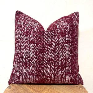 May include: A square decorative pillow with a burgundy and white herringbone pattern. The pillow is made of a textured fabric and is resting on a wooden surface. The pillow is a home decor item.