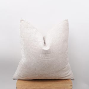 May include: A light beige square pillow with a textured fabric. The pillow is placed on a wooden surface against a white background. The pillow has a soft, inviting appearance, suitable for home decor.