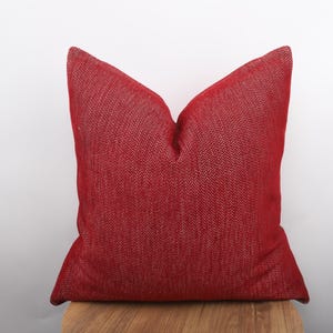 May include: A square, red decorative pillow with a textured weave pattern. The pillow is resting on a wooden surface and has a solid color. The pillow is a home decor item.