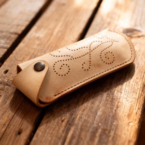 May include: A light tan leather case featuring a decorative pattern of small punched holes in a swirling design. The case is secured with a single dark metal snap button closure on the side flap, maintaining a simple and functional structure.