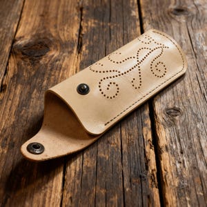 May include: A light beige leather case featuring a decorative pattern of small punched holes in a swirling vine design. The case is secured with a dark metal snap button closure and has a smooth, natural finish on the exterior surface.