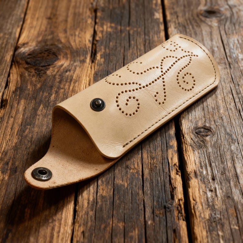 May include: A light beige leather case featuring a decorative pattern of small punched holes in a swirling vine design. The case is secured with a dark metal snap button closure and has a smooth, natural finish on the exterior surface.