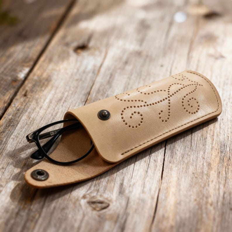 May include: A light tan leather glasses case featuring a decorative perforated swirl pattern on the flap. The case is secured with a dark metal snap button and contains a pair of black-framed eyeglasses partially visible inside the opening.