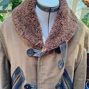 May include: A tan coat with a brown shearling collar and lining. The coat features dark brown leather accents and large, dark buttons. The coat is displayed on a mannequin, showcasing its design and details.