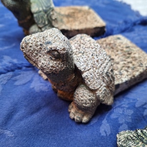 May include: A detailed image of a small, textured turtle figurine with a weathered appearance. The turtle is resting on a rectangular base, with a blue fabric background. The figurine has a detailed head, shell, and feet.