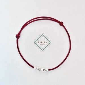 Puede incluir: Un brazalete de cuerda roja con cuentas plateadas. El brazalete está atado con un nudo y tiene un pequeño dije plateado con la palabra "FYPPLEY" en él.
