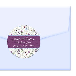 May include: A round, white sticker with a floral design in shades of purple, blue, and white. The sticker has a purple band with the text "Michelle Bolton 123 Main Street Anytown, VA 25456" in white.