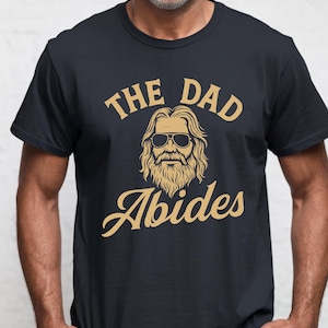 May include: A black short-sleeved t-shirt featuring a tan graphic print of a bearded person with long hair wearing sunglasses. The text above the graphic reads THE DAD and the text below the graphic reads Abides in a stylized cursive font.