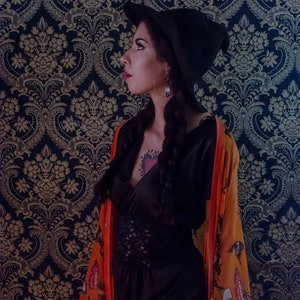 May include: A woman wearing a black witch hat and a black satin dress with a lace trim. She is wearing a long orange kimono with a butterfly pattern. The kimono has a red butterfly on the bottom right corner. The woman has a tattoo of a heart on her chest and a tattoo of a woman on her thigh.