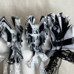 May include: Close-up of three decorative fabric embellishments in black and white. The embellishments feature layered, ruffled fabric with lace accents and ribbon details. The design includes a mix of striped and solid patterns.