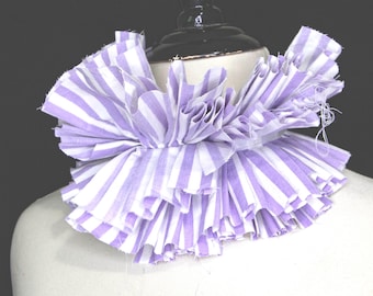 Puede incluir: Un cuello con volantes en un patrón de rayas lavanda y blancas. La tela está fruncida y en capas, creando un efecto voluminoso. El cuello se exhibe en un maniquí.