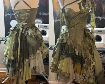 Puede incluir: Un vestido verde único, hecho a mano, con adornos en forma de hojas. El corpiño presenta una espalda de corsé y la falda está superpuesta con varias tonalidades de verdes y beiges. El vestido se exhibe en un maniquí, mostrando su intrincado diseño y texturas.