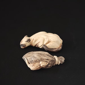 Puede incluir: Dos pequeñas esculturas talladas a mano sobre un fondo negro. Una es un oso beige claro, la otra un saco beige con un patrón texturizado. El oso tiene acentos marrón oscuro en la cara y las patas. El saco está atado en la parte superior.