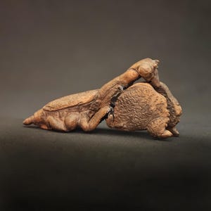 May include: A brown, textured sculpture of a praying mantis in a resting position. The figure features detailed, carved-like surfaces with a dark, glossy accent along the midsection where the body segments meet, showcasing a realistic insect form.