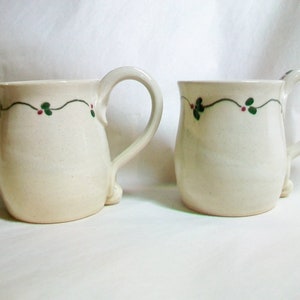 May include: Two white ceramic mugs with green and red holly designs. The mugs have a rounded shape and a handle.
