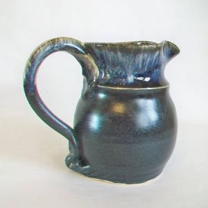 May include: A handmade ceramic pitcher with a dark blue and black glaze. The pitcher has a rounded body, a curved handle, and a spout. The rim of the pitcher is a lighter blue color. The pitcher is on a white background.