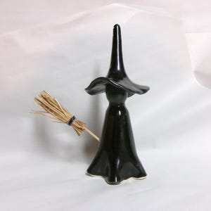 May include: A black ceramic witch figurine with a wide-brimmed hat and a long dress. The figurine holds a small broom made of straw. The witch is a Halloween or fall-themed decorative item.
