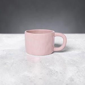 May include: A light pink ceramic mug with a smooth, rounded silhouette and a thick, curved handle. The mug features a subtle, organic texture on its exterior surface, giving it a slightly uneven, handcrafted appearance with a soft matte finish.