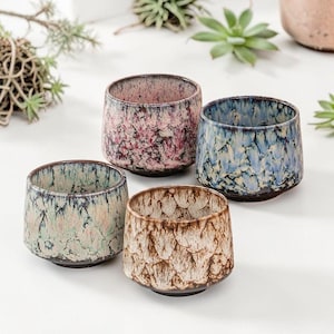 May include: Four ceramic cups with textured surfaces in shades of pink, blue, green, and brown. The cups have a speckled glaze and a dark rim and base. The cups are arranged on a white surface, suggesting a decorative or functional use.