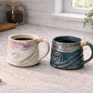 May include: Two ceramic mugs filled with coffee. One is off-white with a swirl design and a rope-wrapped handle. The other is teal with a similar design and handle. Both mugs contain dark coffee. The mugs are on a countertop.