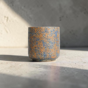 May include: A cylindrical ceramic cup featuring a textured, mottled surface with a blend of vibrant orange and deep blue speckles. The vessel has a straight-sided profile with a flat base and a smooth, rounded rim, showcasing a distinct granular finish.
