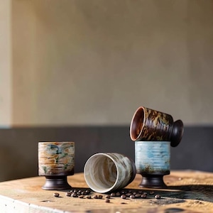May include: Four ceramic cups with pedestal bases and marbled glaze patterns in shades of brown, blue, and cream. One cup is positioned on its side, while the others stand upright. Scattered dark roasted coffee beans surround the cup bases.