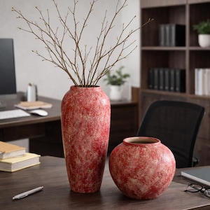 Könnte beinhalten: Zwei rot-cremefarbene Keramikvasen, eine hoch und zylindrisch, die andere rund, stehen auf einem dunklen Holzschreibtisch. Die hohe Vase enthält kahle Zweige. Ein Stift und Bücher liegen ebenfalls auf dem Schreibtisch. Der Hintergrund umfasst einen Schreibtisch und Regale.