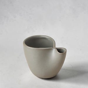 Może przedstawiać: Matowy, jasnoszary ceramiczny dzbanek na mleko lub mały dzbanek o zaokrąglonym, asymetrycznym korpusie z wyraźnym, zintegrowanym dzióbkiem. Wnętrze naczynia ma nieco ciemniejszy odcień szarości, a zewnętrzna powierzchnia jest gładka i jednolita.