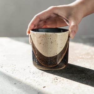 Könnte beinhalten: Ein Keramikbecher mit zweifarbigem Design, bestehend aus einem gesprenkelten cremefarbenen oberen Bereich und einem dunkelbraunen, texturierten unteren Bereich. Der Becher hat eine zylindrische Form mit glattem Rand und geschwungenem Übergang.
