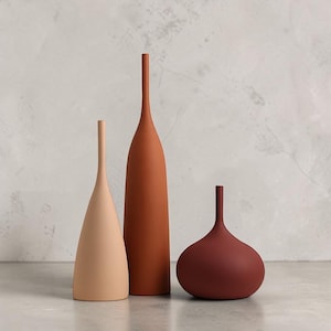 Può includere: Tre vasi in ceramica di varie forme e colori. Il vaso più alto è arancione tenue, il vaso centrale è color pesca chiaro e il vaso più corto è bordeaux intenso. Tutti hanno una finitura opaca e sono esposti su una superficie neutra.