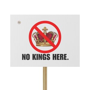 May include: A white sign with the words "NO KINGS HERE." A red circle with a line through it surrounds a gold and red crown. The sign is mounted on a wooden pole.