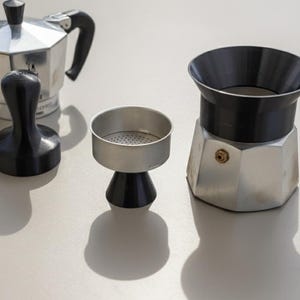 Op de afbeelding: Een verzameling espressomakers, waaronder een zilveren espressopot, een zwarte tamper en een filtermand. De espressopot heeft een zwarte handgreep en bovenkant. De items zijn gerangschikt op een lichtgekleurd oppervlak.