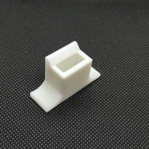 Puede incluir: Objeto blanco de plástico impreso en 3D con una abertura rectangular. El objeto tiene una base curva y una parte superior plana.