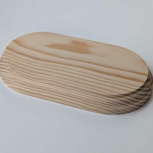 Puede incluir: Una bandeja ovalada de madera natural con un acabado liso de color marrón claro. La bandeja está hecha de madera maciza y tiene un borde ligeramente redondeado.