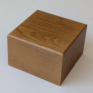 Può includere: Una scatola quadrata in legno massello con una finitura color legno chiaro. La scatola ha una superficie liscia e bordi arrotondati.