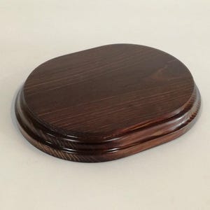 Könnte beinhalten: Ein ovaler Sockel aus dunkelbraunem Holz mit glatter, polierter Oberfläche. Der Sockel hat einen doppelten Rand.