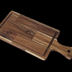 May include: A rectangular wooden cutting board with a handle. The board is made of various wood pieces, creating a mosaic pattern. A groove runs along the edge of the cutting surface. The handle has a hole for hanging.